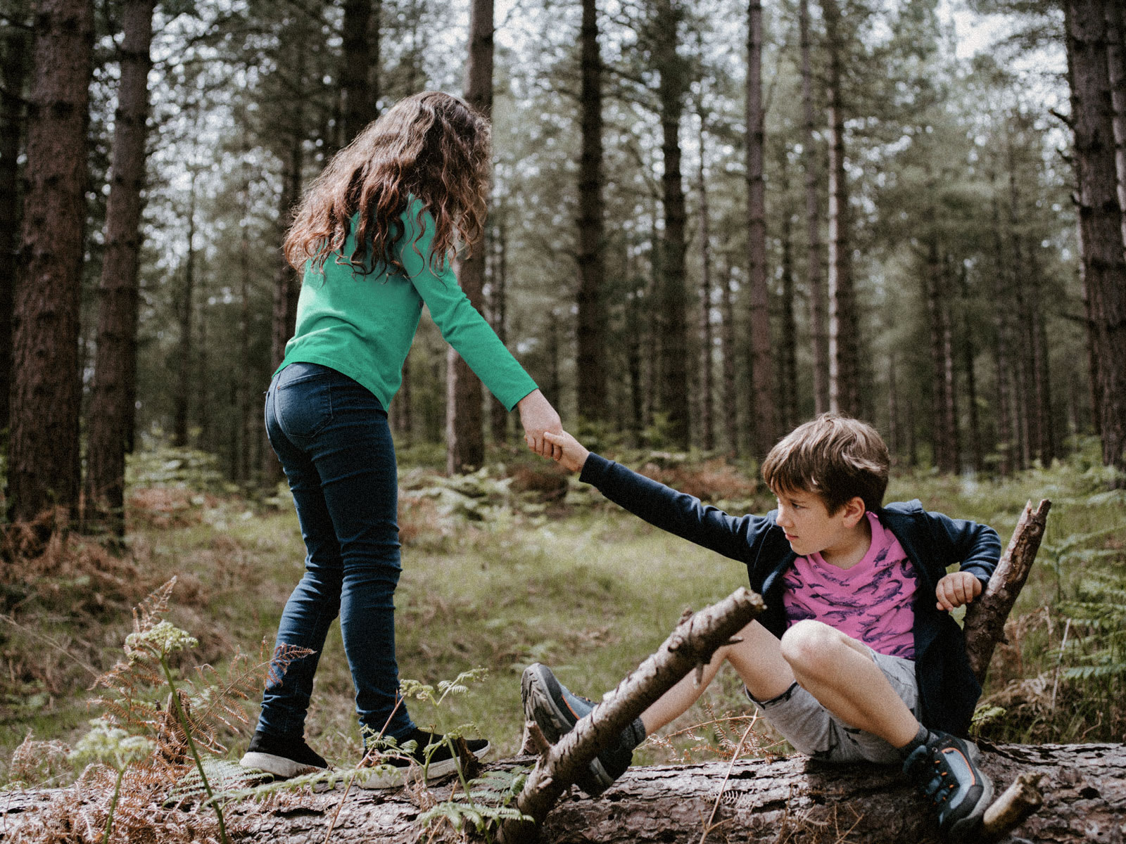Zwei Kinder spielen gemeinsam auf einem gefällten Baumstamm im Wald. Wir sehen, wie eines der beiden Kinder dem anderen nach einem Fall wieder aufhilft.